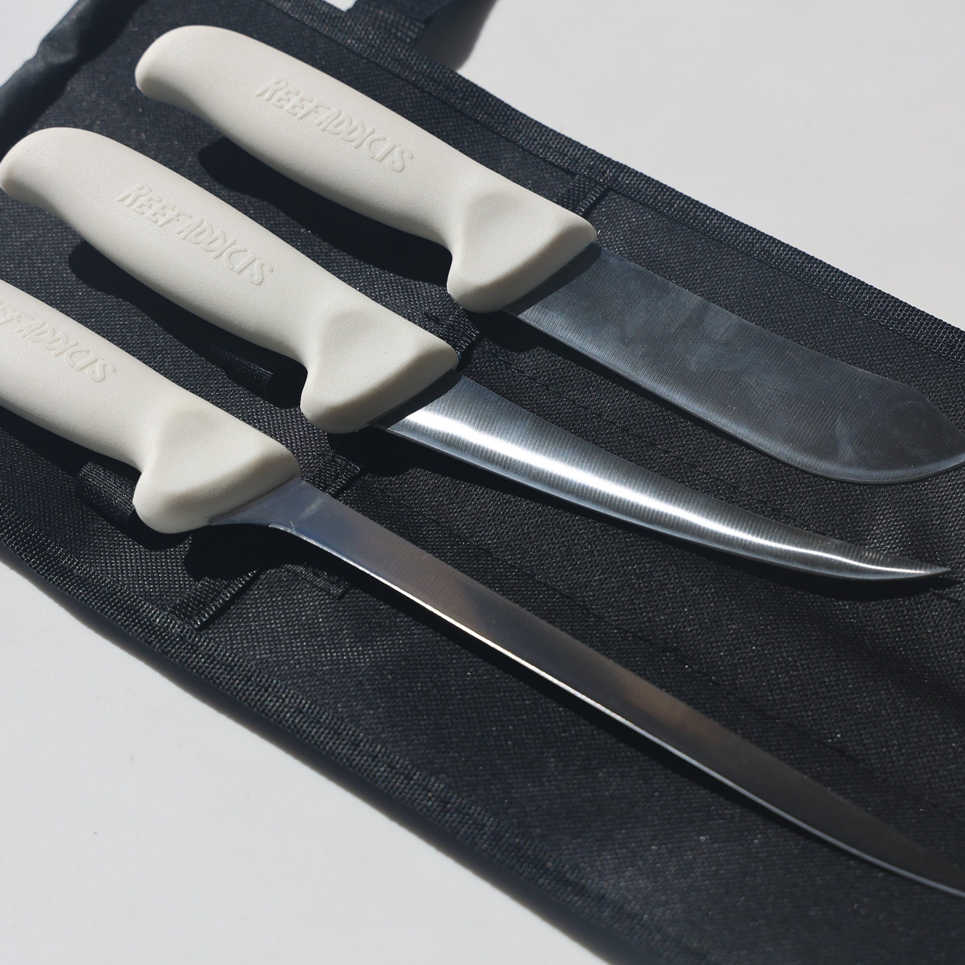 A set of three fishing knives with white handles laid out on a black surface.
